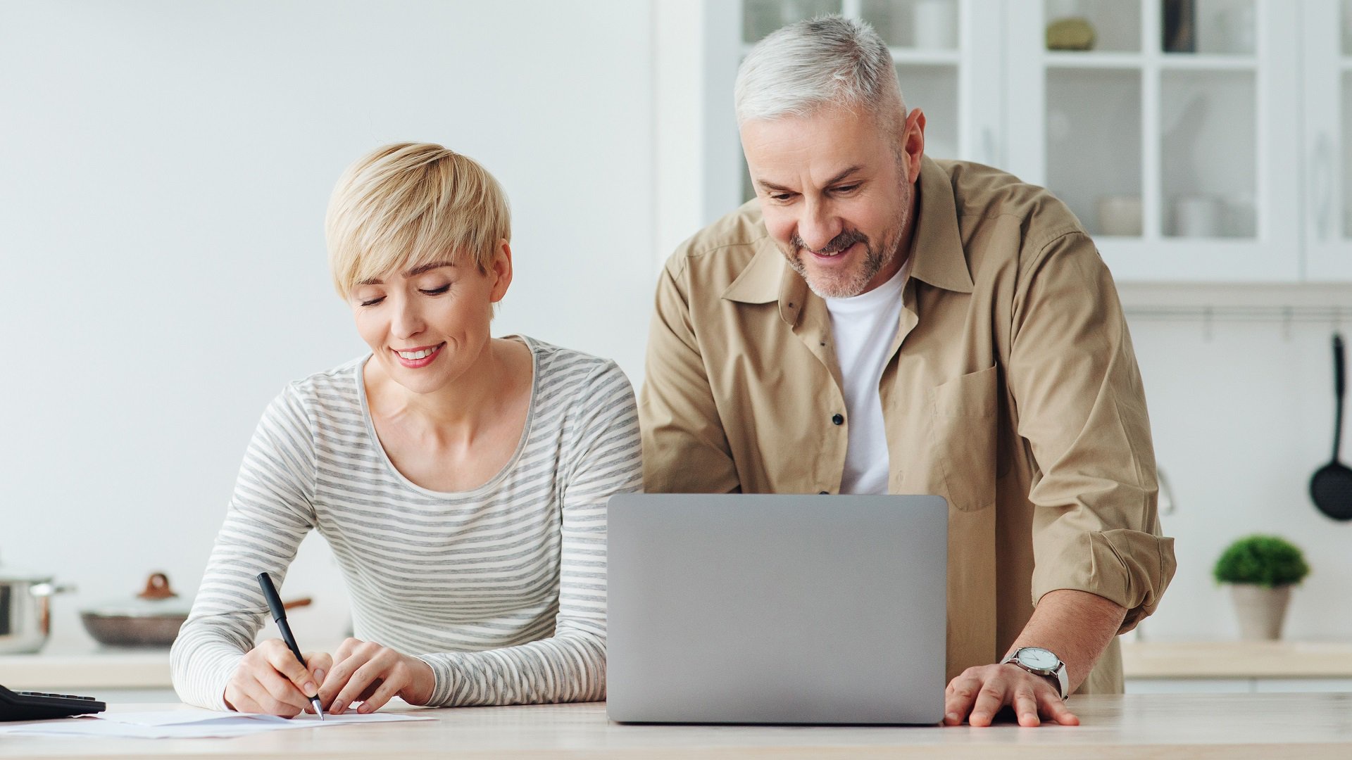 Discover a smiling senior couple checking their finances and paying bills inside their kitchen with a laptop.