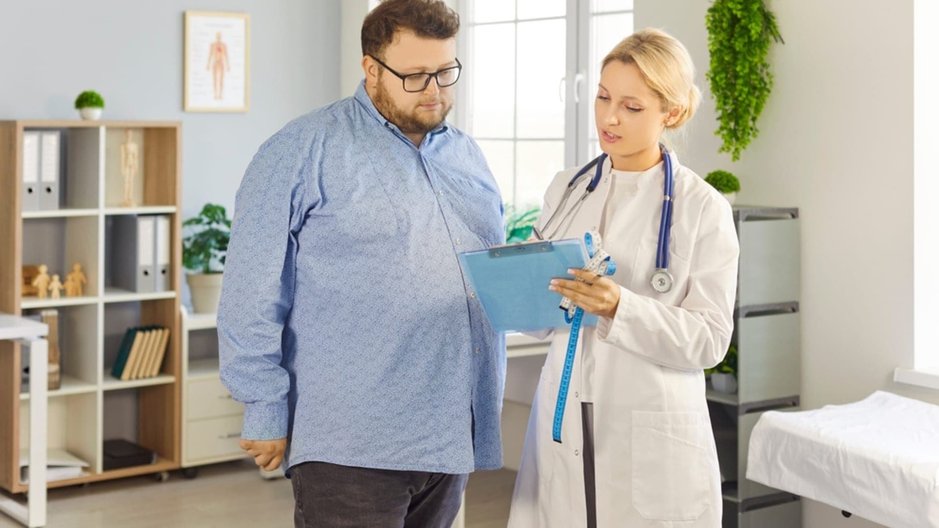 A doctor holds a clipboard and a tape measure while talking with an overweight man in a clinic. Credit: Shutterstock
