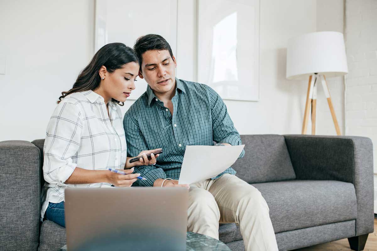 A man and a woman sit on a gray couch in a living room. They are both looking at a piece of paper the man is holding. The woman holds a pen and smartphone. A laptop is open on a table in front of them. There is a lamp in the background.