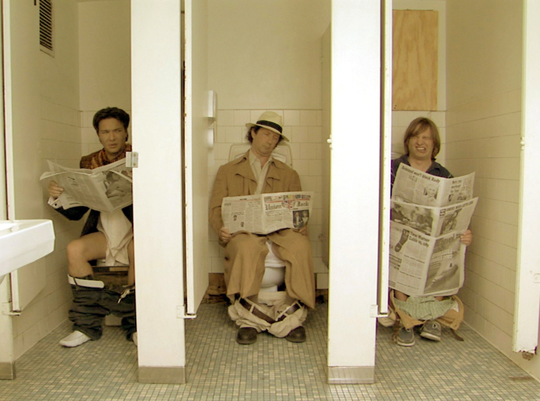 Three people sit in adjacent bathroom stalls, reading newspapers with their pants around their ankles. Appears humorous or staged