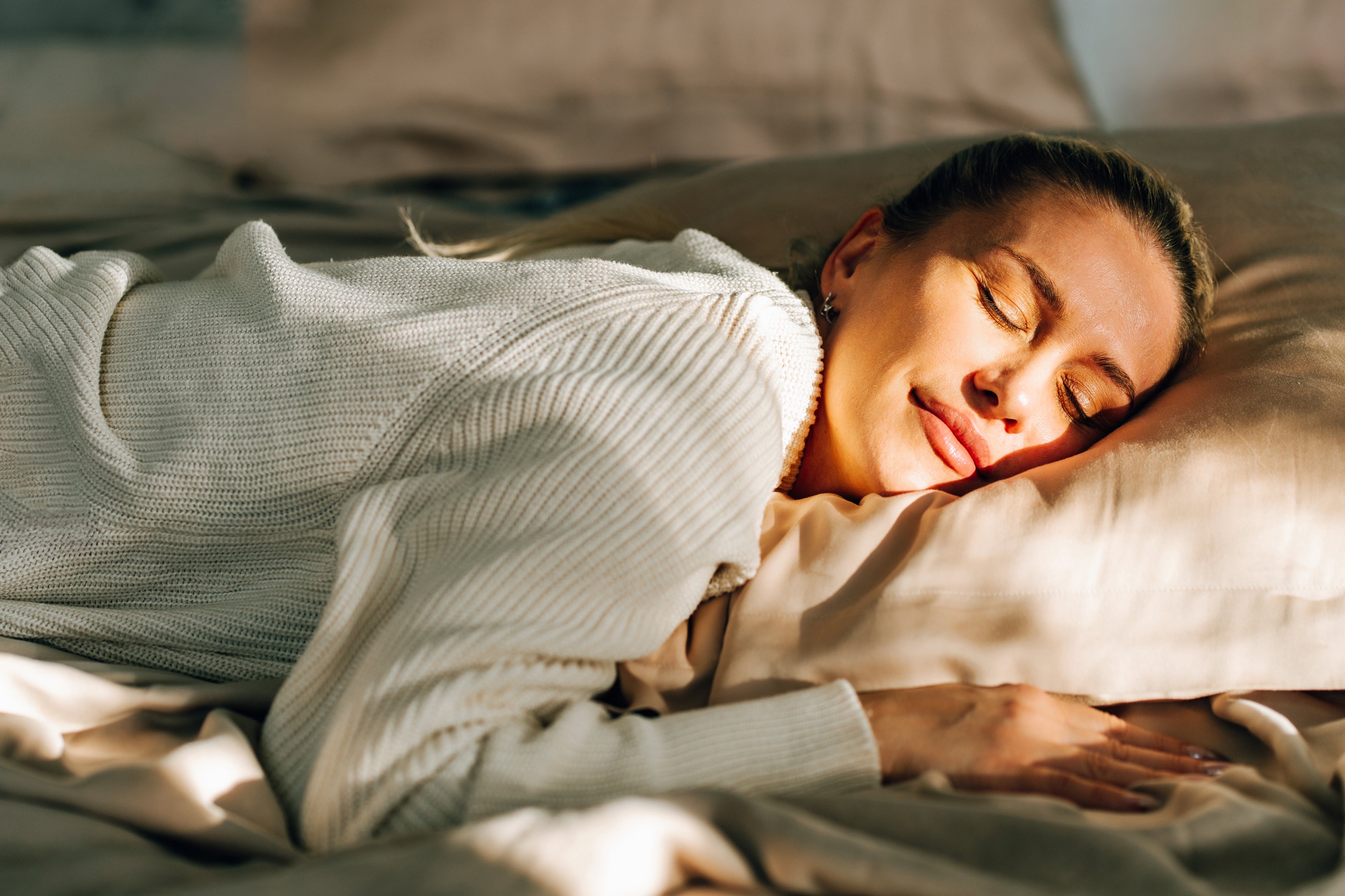 A person peacefully sleeps on a bed, wearing a cozy sweater, with sunlight softly illuminating their face