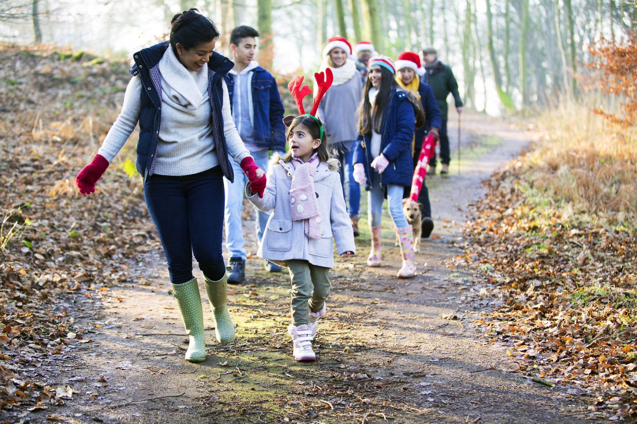 A woman holding a young girl’s hand leads a group of people wearing winter clothes and Santa hats on a walk along a wooded trail.