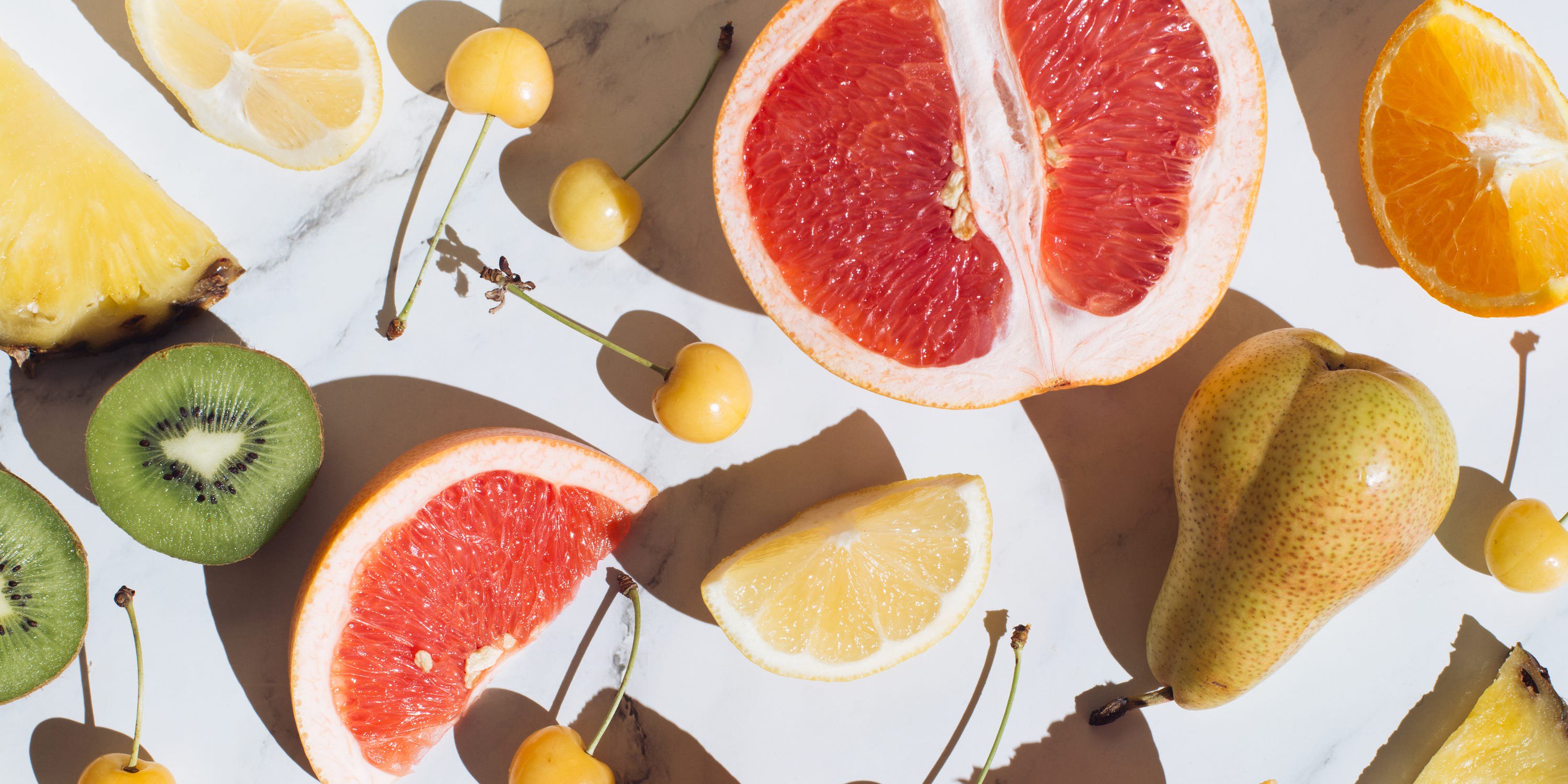 colorful summer fruits on white marble table. fresh raw food concept. creative healthy food background. orange, kiwi, grapefruit, pineapple, pear. flat lay, top view. long shadow and refraction.
