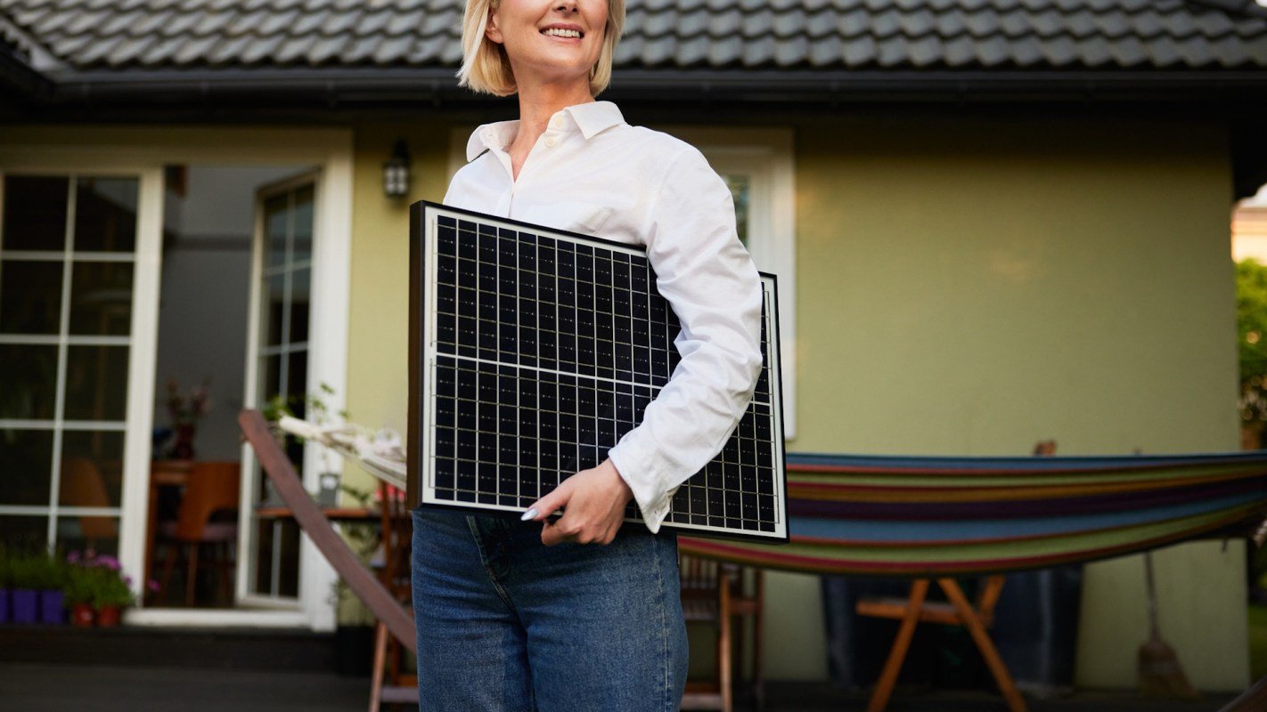 A smiling woman holds a solar panel outside a house with a hammock in the background.