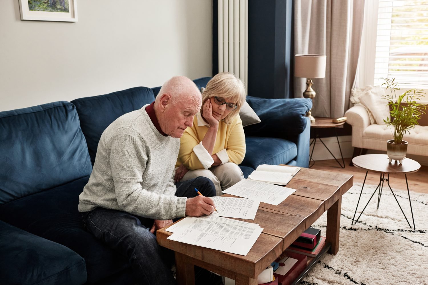 An elderly man in a light grey sweater and dark jeans sits on a navy blue sofa, writing on documents placed on a rustic wooden coffee table. An elderly woman with blonde hair and glasses, wearing a yellow sweater, sits beside him, looking thoughtfully at the papers with her chin resting on her hand. In the background, a cozy living room with another armchair, lamp, and plant is visible.