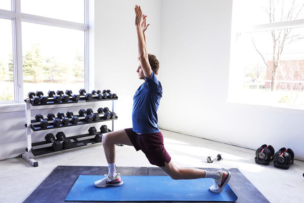 Male exercising in a lunge position with arms raised, in a bright gym with dumbbells in the background.