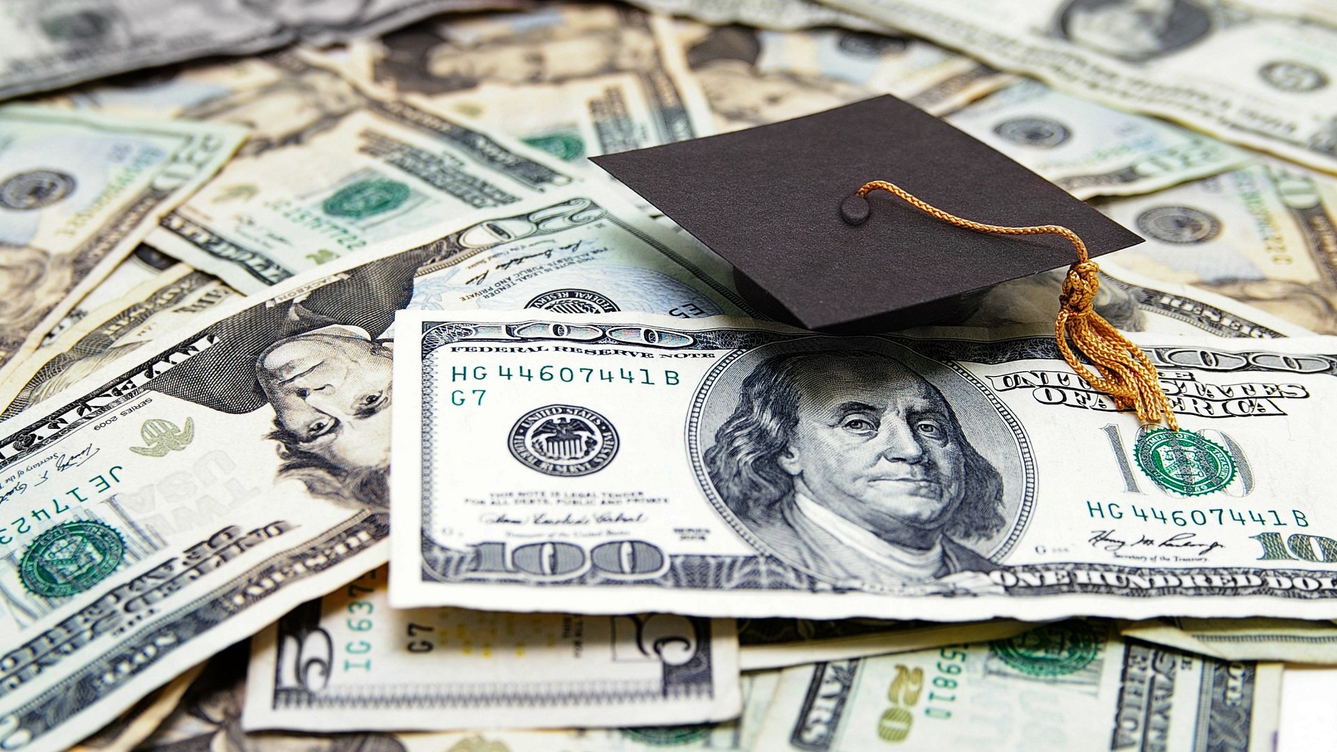 Discover a graduation cap or mortarboard is sitting on top of a pile of cash money to indicate a student loan concept