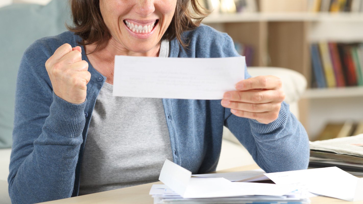 A woman holds a letter while sitting at a table with scattered papers, pumping her fist.