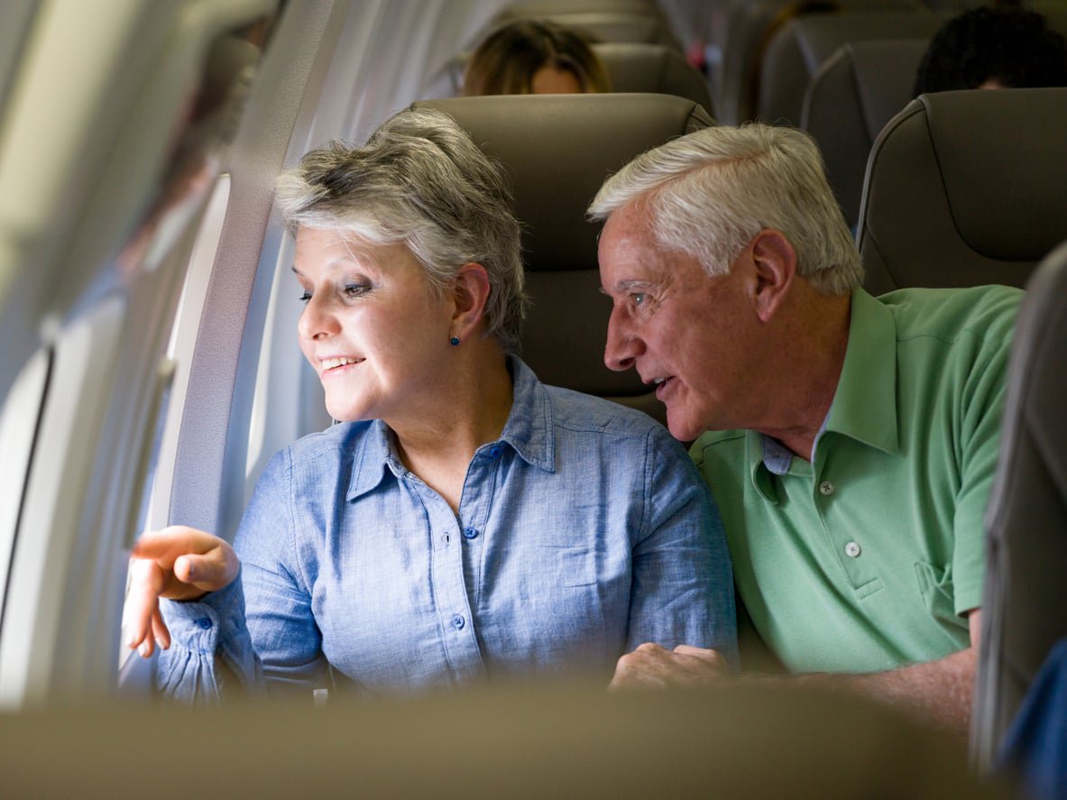 An older couple sitting in airplane seats, with the woman pointing out the window and the man looking in the same direction.