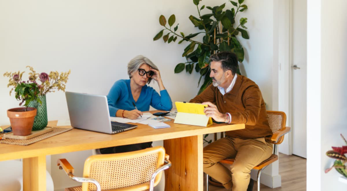 Middle aged couple sit at dining room table together, shooting concerned looks at each other while going over paperwork.