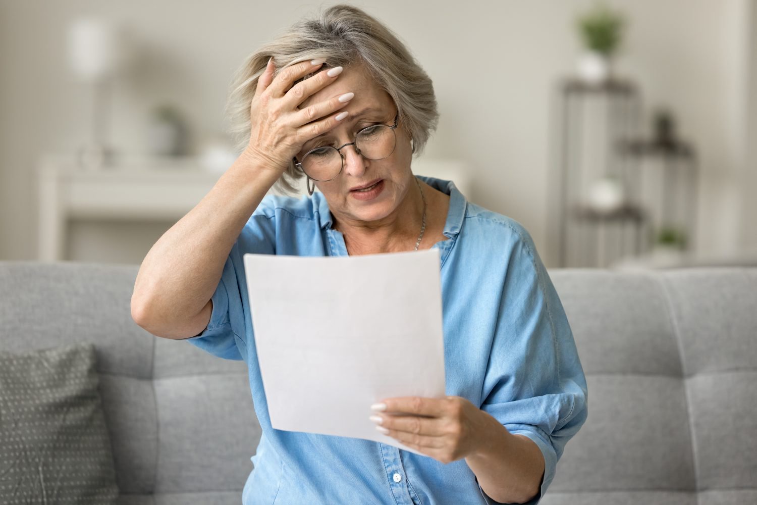 A senior woman with glasses and short, light gray hair sits on a gray couch, holding a white paper in her hands. She is wearing a light blue denim-style shirt. Her left hand is pressed against her forehead, and her expression shows distress and confusion as she looks down at the document.