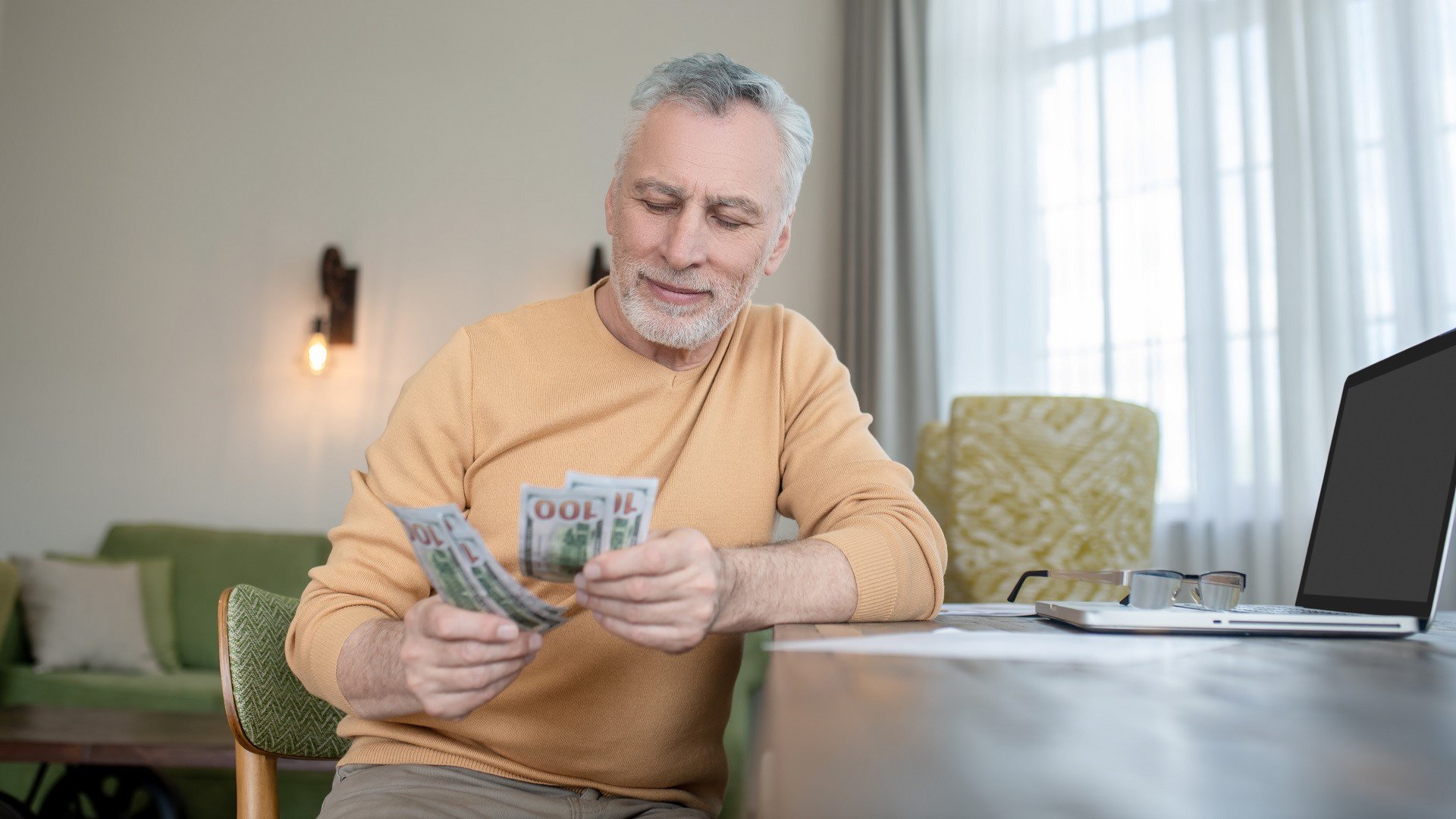 Discover a smiling gray-haired man sitting at his table and counting hundred-dollar bills inside his home.