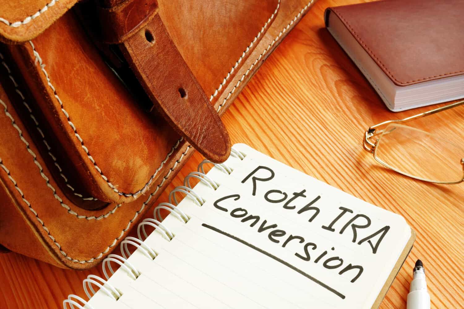 A top-down view of a wooden desk shows a brown leather satchel on the left. In the center, a white spiral notebook is open, with 'Roth IRA Conversion' written neatly in black marker. To the right, a small brown bound book and a pair of gold-rimmed reading glasses are placed next to a black marker.
