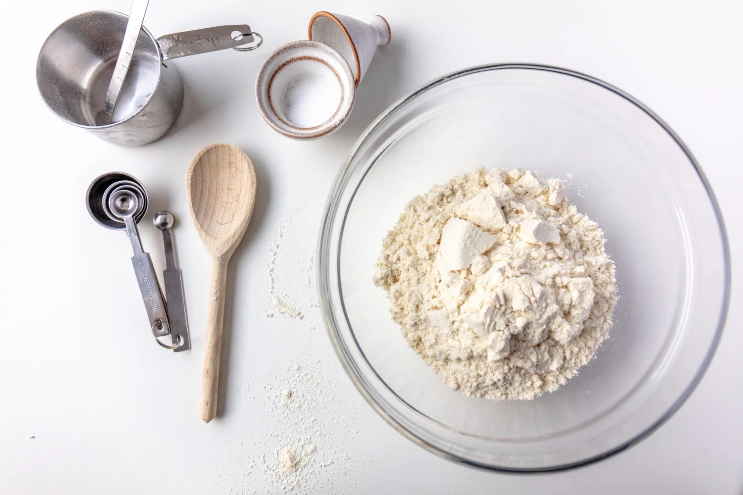 A bowl of masa harina next to measuring tools and a wooden spoon on a table