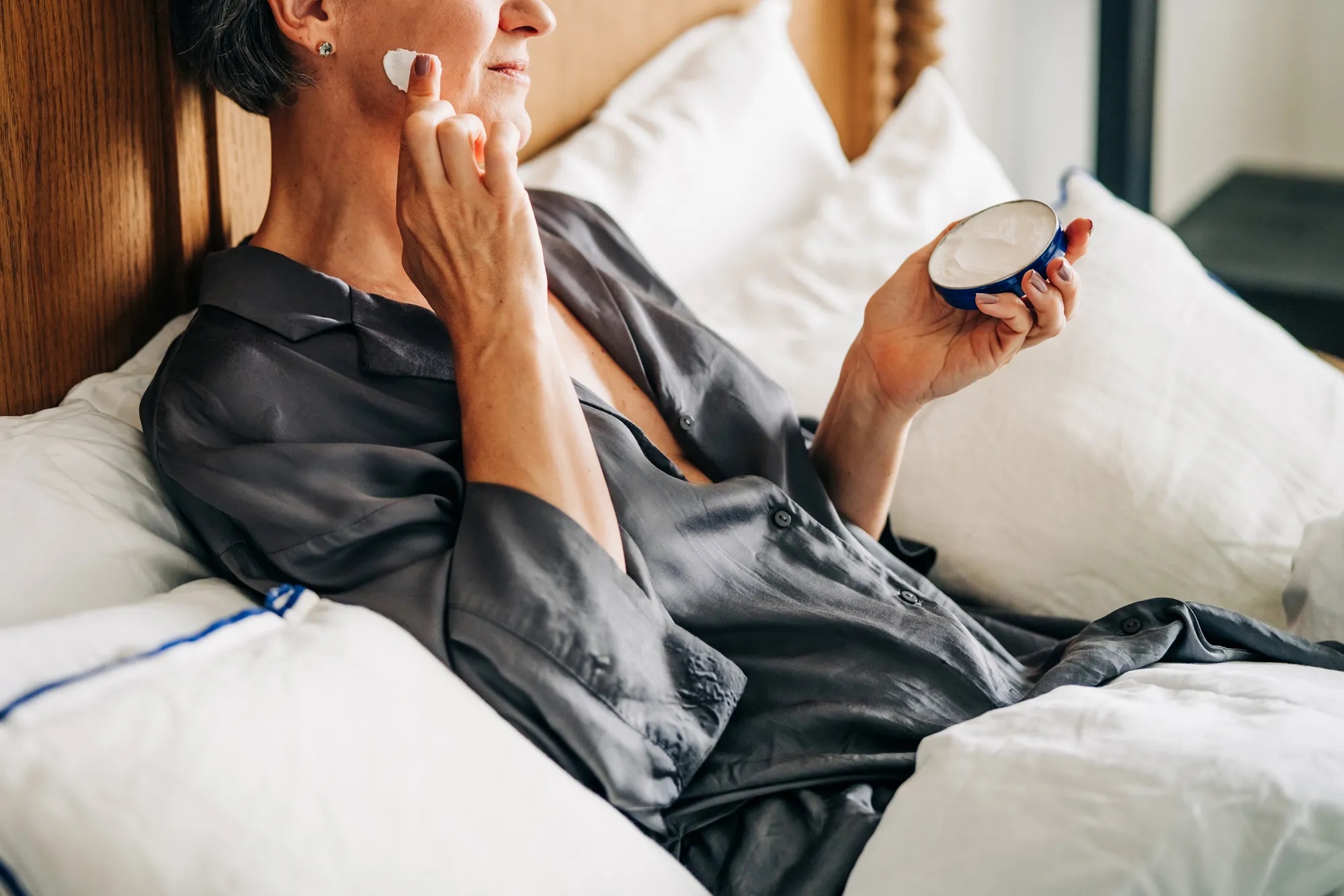 A woman sits on a bed and applies moisturizer cream.