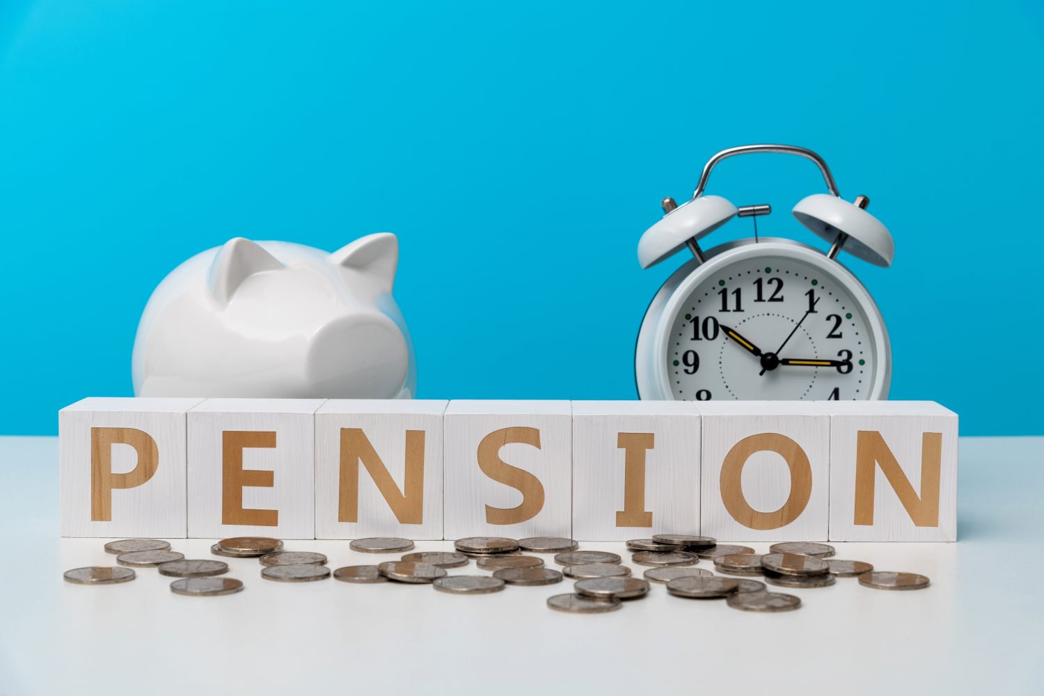 A white piggy bank, a white alarm clock set to approximately 3:45, and scattered silver coins sit on a white surface against a light blue background. In the foreground, white wooden blocks spell out the word 'PENSION' in gold letters, with coins directly in front of them.