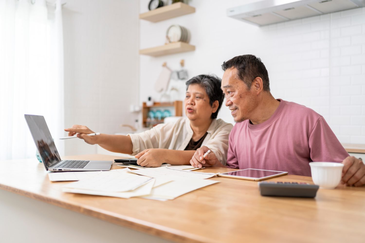 An older Asian couple sits at a bright wooden counter in a modern kitchen. The woman on the left points at a laptop screen, while the man on the right looks at it intently, holding a white pen. Several papers, a tablet, a calculator, and a white coffee cup are scattered across the counter.