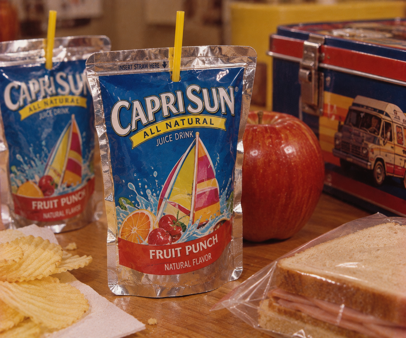 Capri Sun juice pouches, an apple, a sandwich in a plastic bag, potato chips, and a colorful lunchbox recall 1980s grocery store staples arranged on a table.