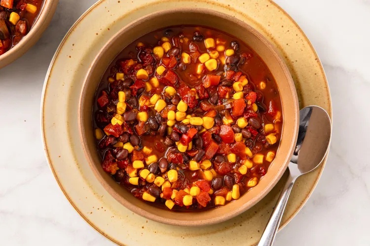 A bowl of hearty soup containing corn black beans and tomatoes with a spoon on the side