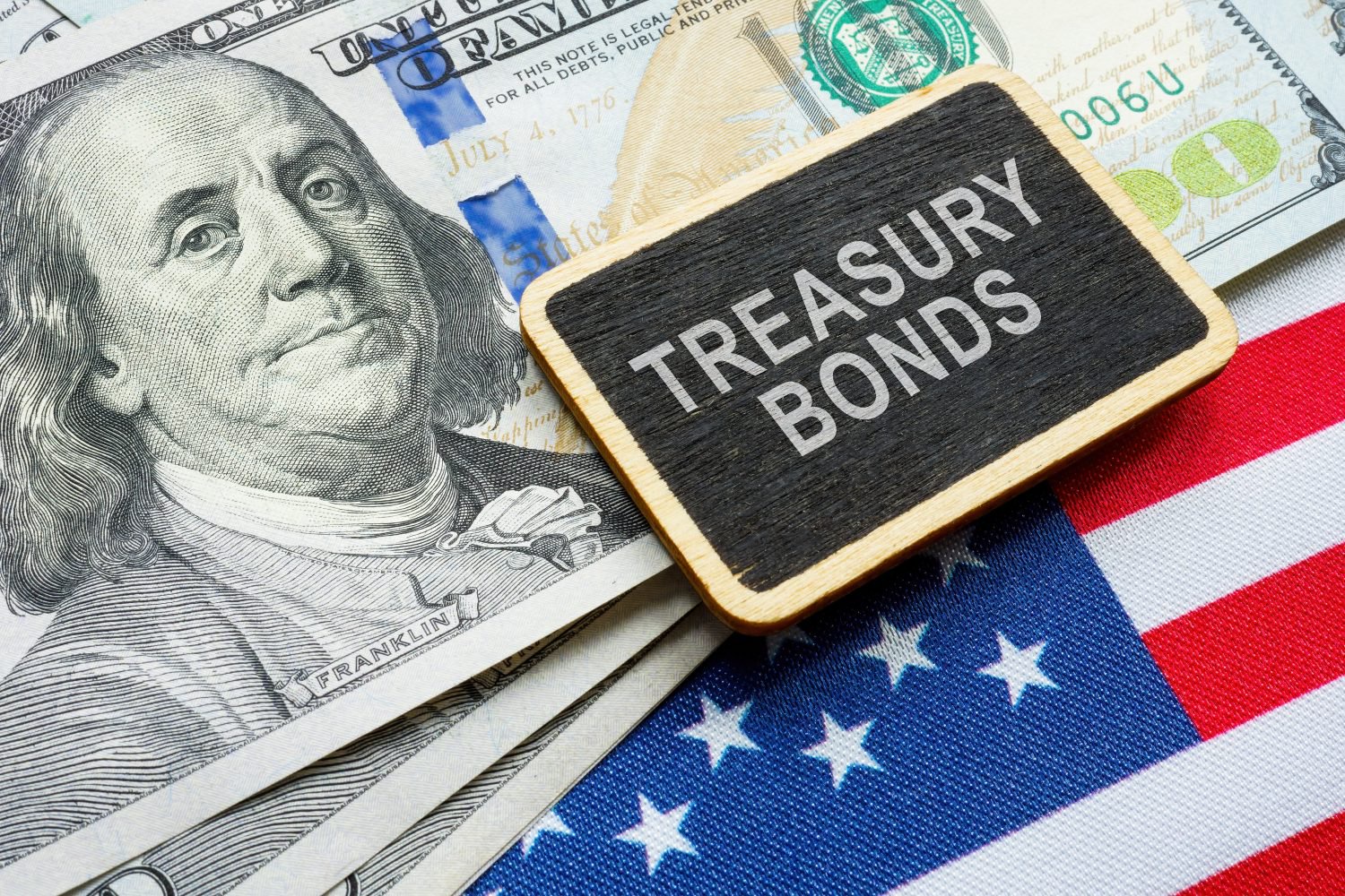 Close-up of a stack of U.S. hundred-dollar bills featuring Benjamin Franklin, partially covered by an American flag. A small black chalkboard sign with a wooden frame rests on top, displaying the words 'TREASURY BONDS' in white capital letters.