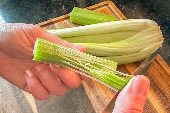 A hand uses a knife to remove celery strings from a stalk over a wooden cutting board with more celery in the background.