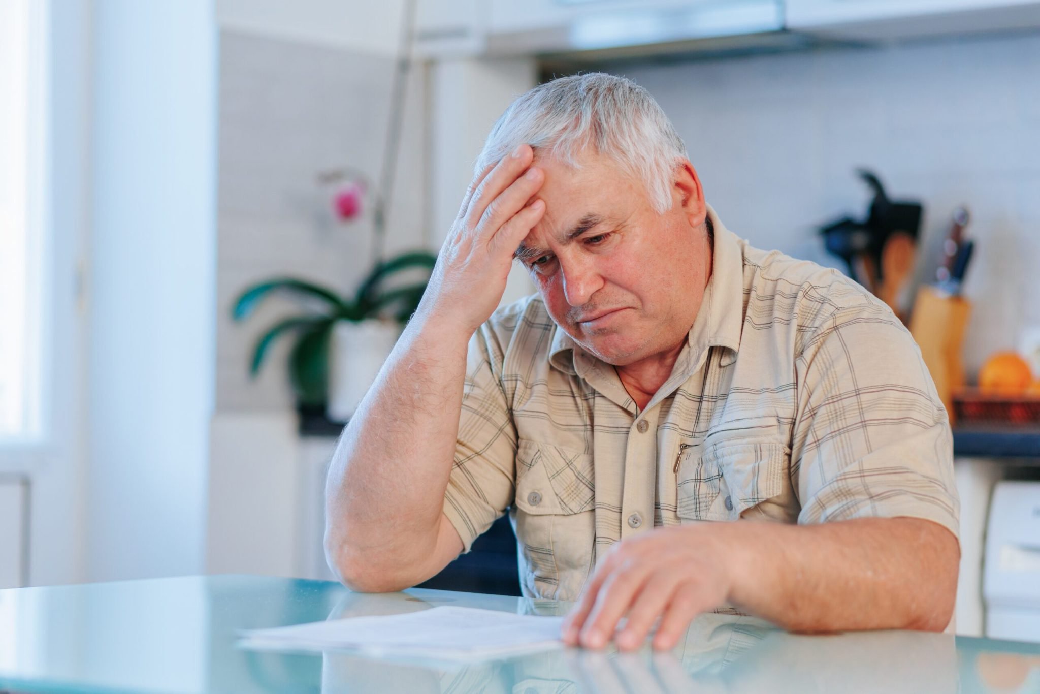 A somber-looking older man with white hair sits at a light-colored table, holding his right hand to his forehead. He is wearing a plaid button-down shirt and looking down at white papers on the table in front of him. The background is a brightly lit, blurred kitchen or dining area with a window on the left and a plant and kitchen utensils visible on the right.
