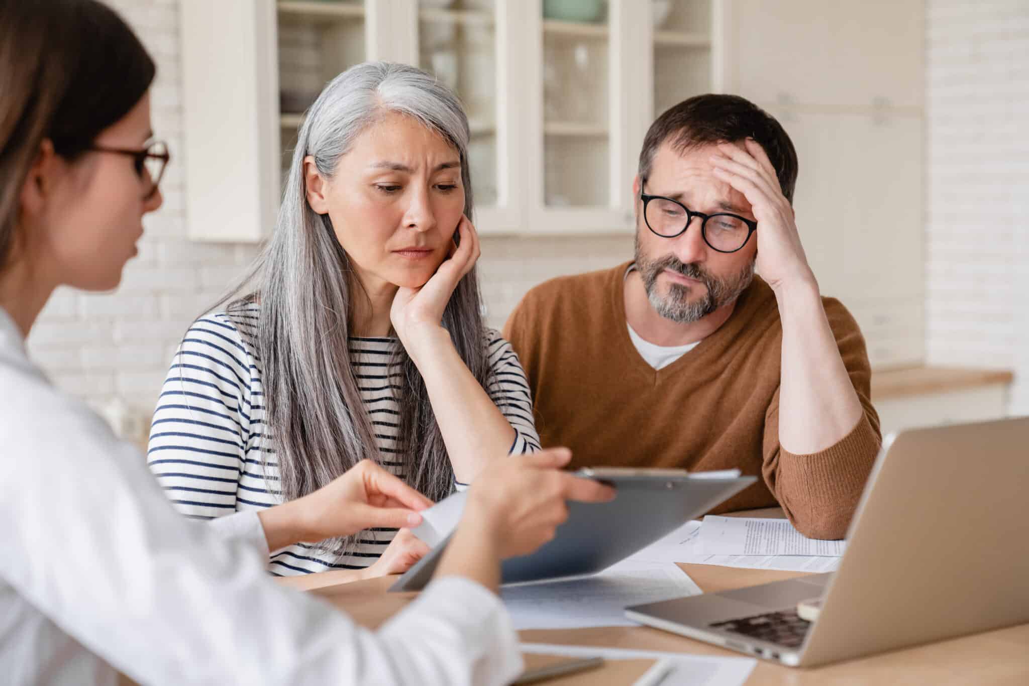 Three people are seated at a table. On the left, a woman in a white coat holds a clipboard, pointing to documents. In the center, an Asian woman with long grey hair and a striped shirt rests her chin on her hand, looking concerned. To her right, a bearded man in glasses and a brown sweater holds his head in his hand, appearing stressed. A laptop and other documents are visible on the table.