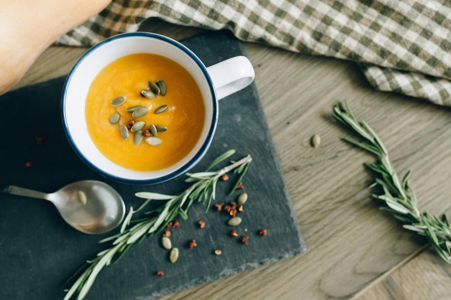 Image of a mug of pumpkin soup, topped with pepita pumpkin seeds with a spoon and sprigs of rosemary beside it.