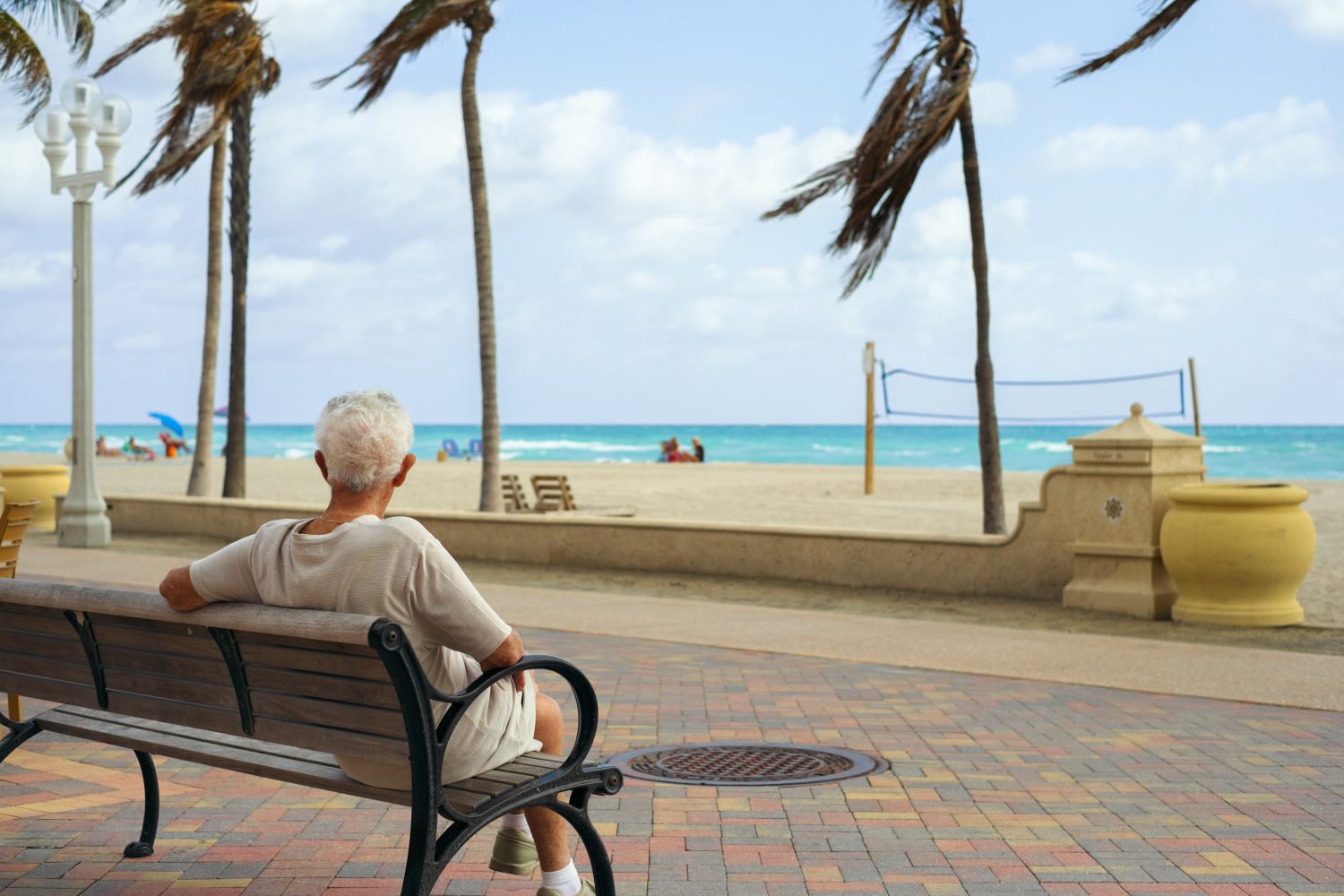 An elderly person with white hair sits on a dark brown and black park bench, facing away from the camera, looking out at a sunny beach and the turquoise ocean. In the background, there are several palm trees, a white sand beach with a blue volleyball net, and a few distant beachgoers. The foreground features a paved walkway with multi-colored bricks and a manhole cover.