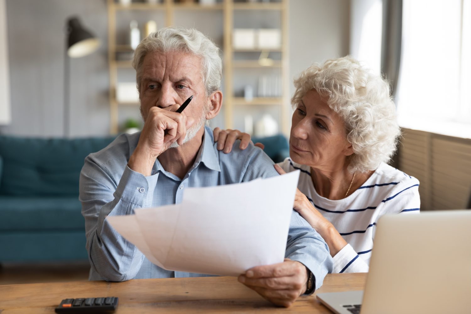 A white-haired older man with a beard, wearing a light blue button-up shirt, sits at a wooden table, holding white papers and looking down with a worried expression. His left hand is raised to his chin, holding a pen. Next to him, a white-haired older woman with curly hair, wearing a white and blue striped shirt, places her right hand on his left shoulder, looking at him with a concerned expression. A black calculator and part of a silver laptop are on the table. The background shows a living room with a teal sofa and shelves.