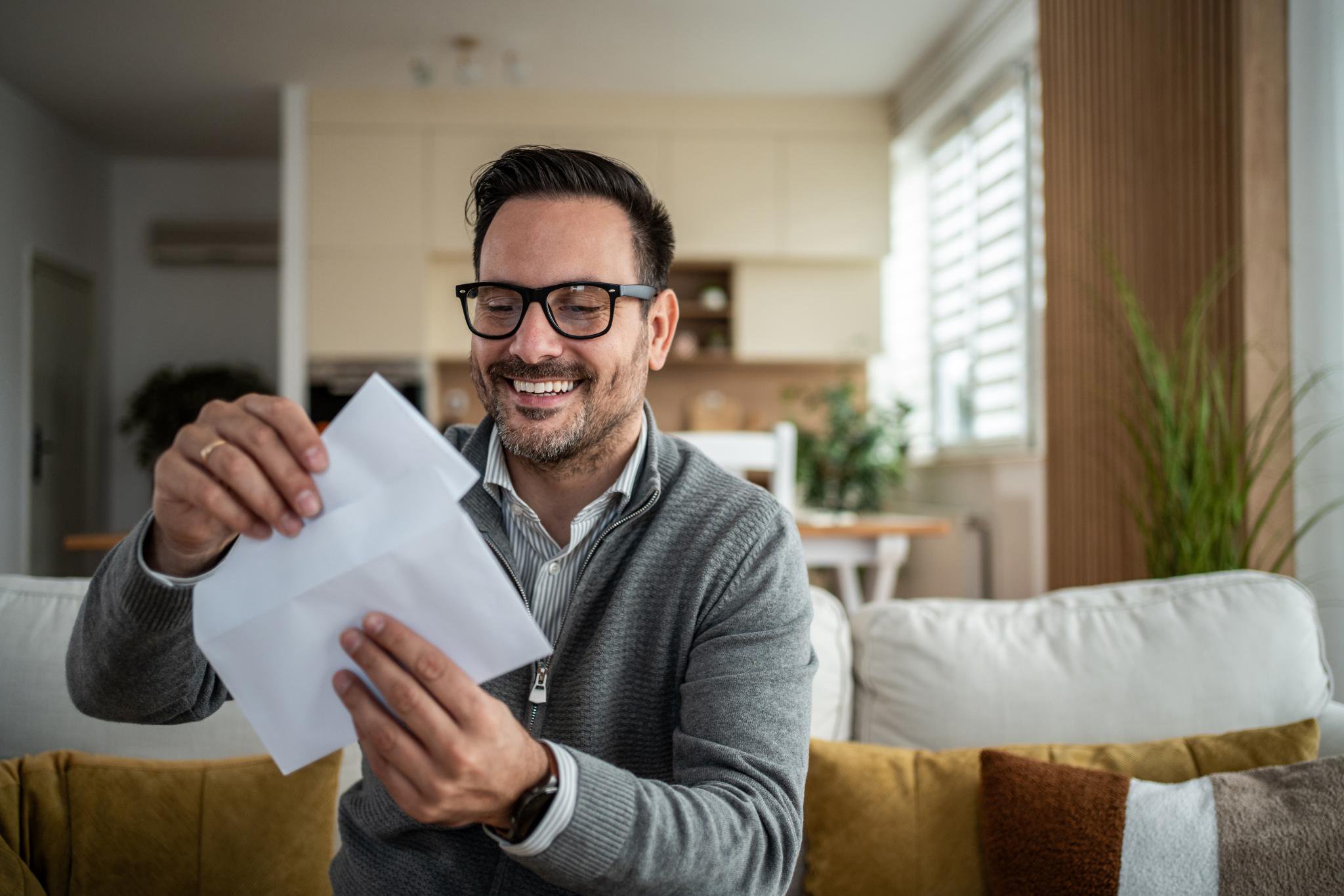 A smiling man wearing glasses sits on a sofa while opening an envelope with a letter.