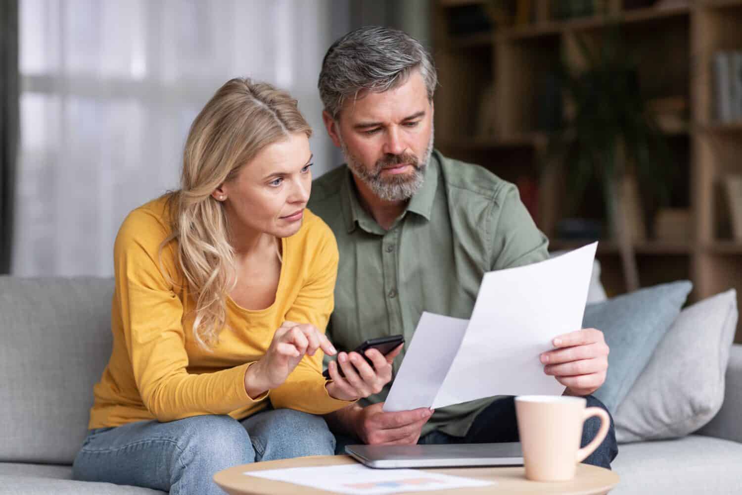 A middle-aged man with graying hair and a beard, wearing an olive green shirt, sits on a grey sofa next to a blonde woman in a yellow long-sleeved shirt and blue jeans. They are both looking down at white papers held by the man, while the woman points to a black smartphone he holds. A silver laptop and a light beige mug are visible on a wooden coffee table in the foreground, with a blurred bookshelf in the background.
