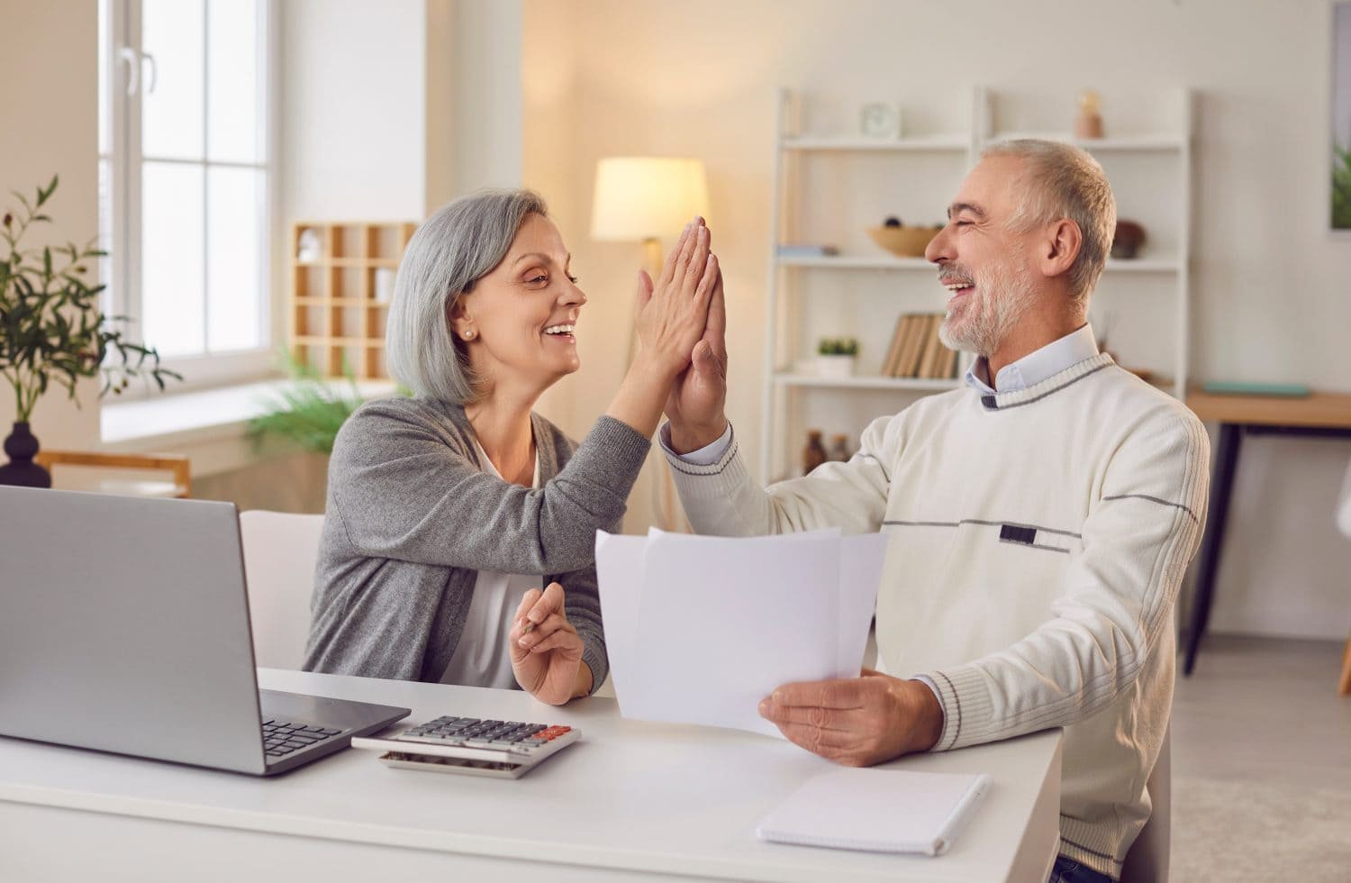 A smiling older woman with grey hair and an older man with a grey beard high-five across a white desk. On the desk are a silver laptop, a calculator, and white papers. The couple appears happy and engaged in financial planning in a bright living room setting.