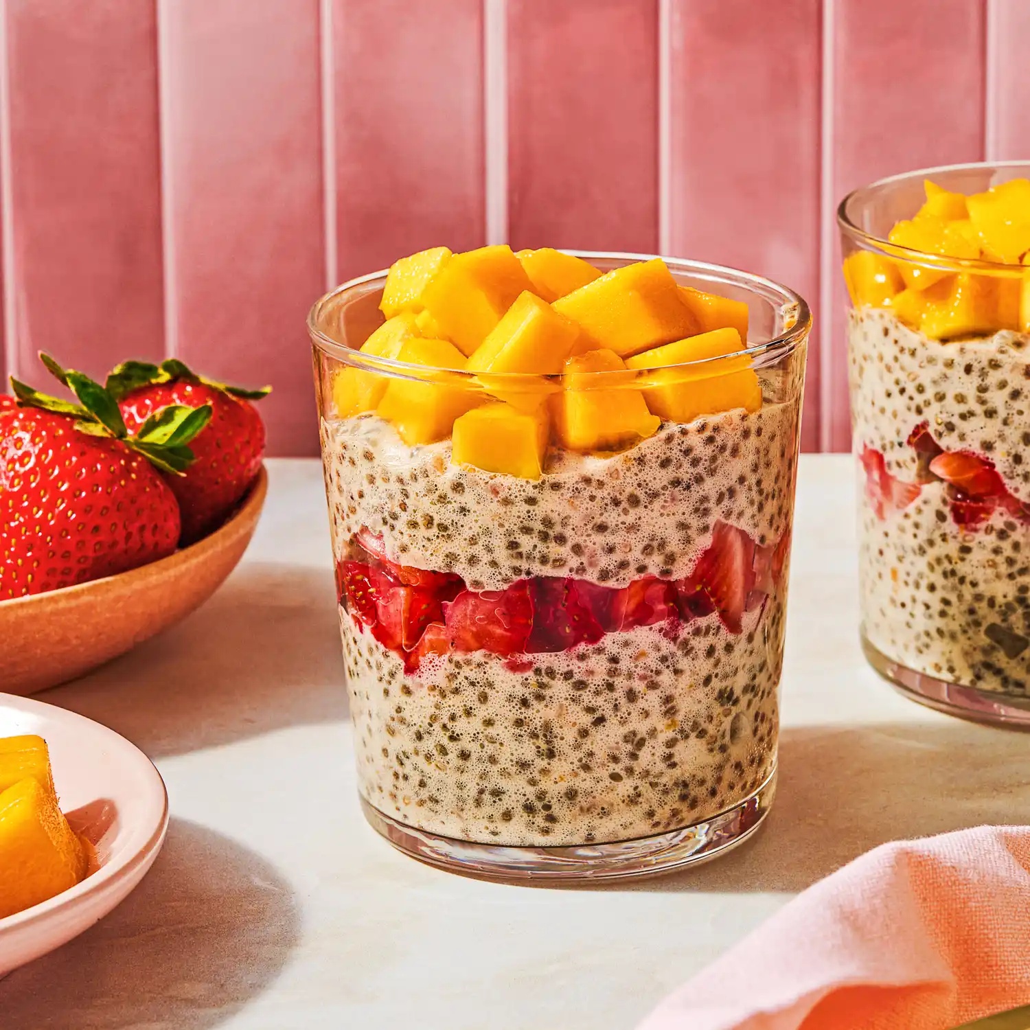 Chia pudding in a glass topped with mango and strawberries with extra fruit nearby