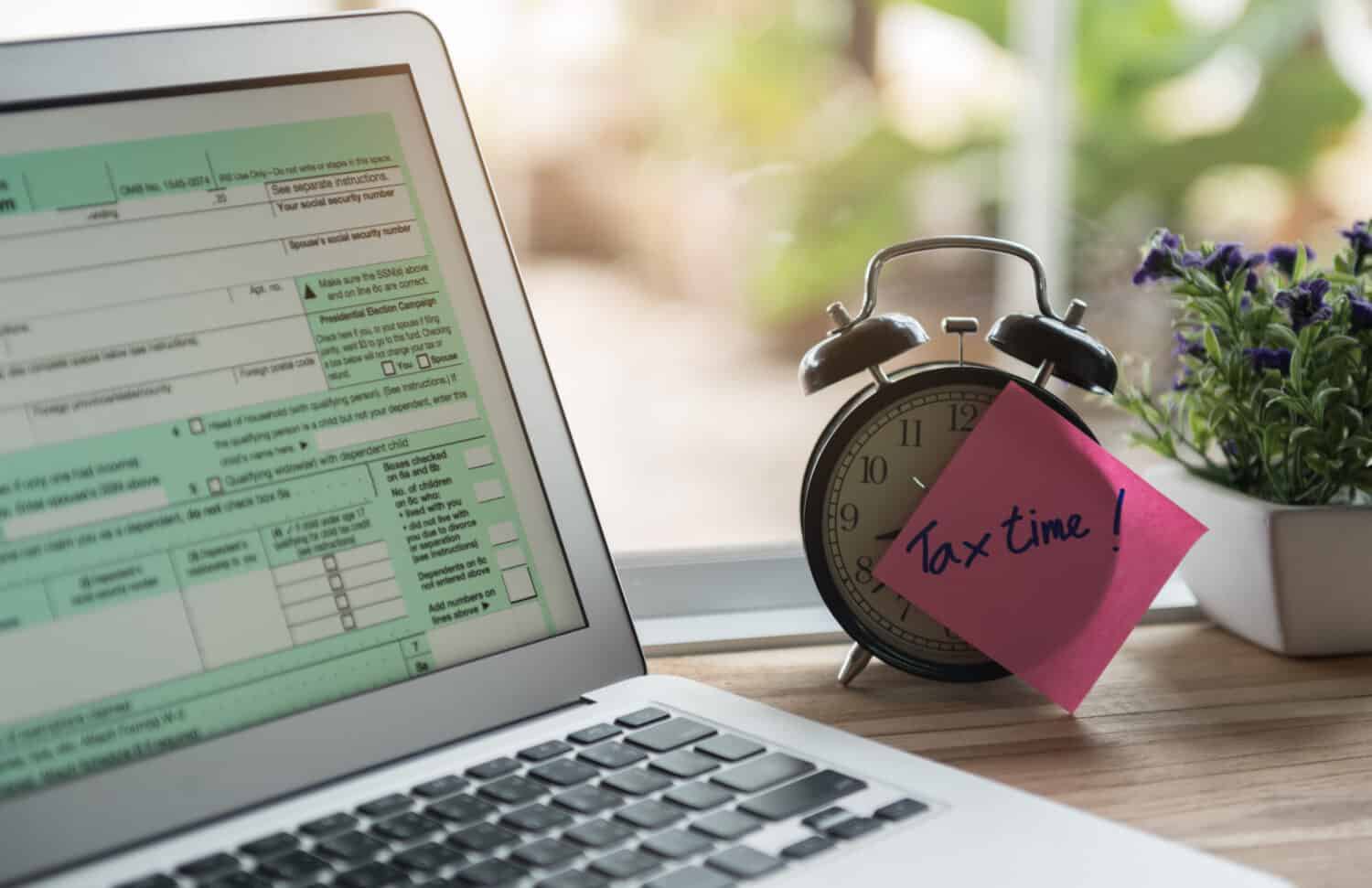 A close-up shot shows an open silver laptop displaying a green-hued IRS tax form on its screen, positioned on a light wooden desk. To the right of the laptop is a vintage-style black alarm clock with white numbers, on which a bright pink square sticky note reads 'Tax time!' in blue handwritten script. A potted plant with small purple flowers is visible in the background, suggesting an indoor setting.