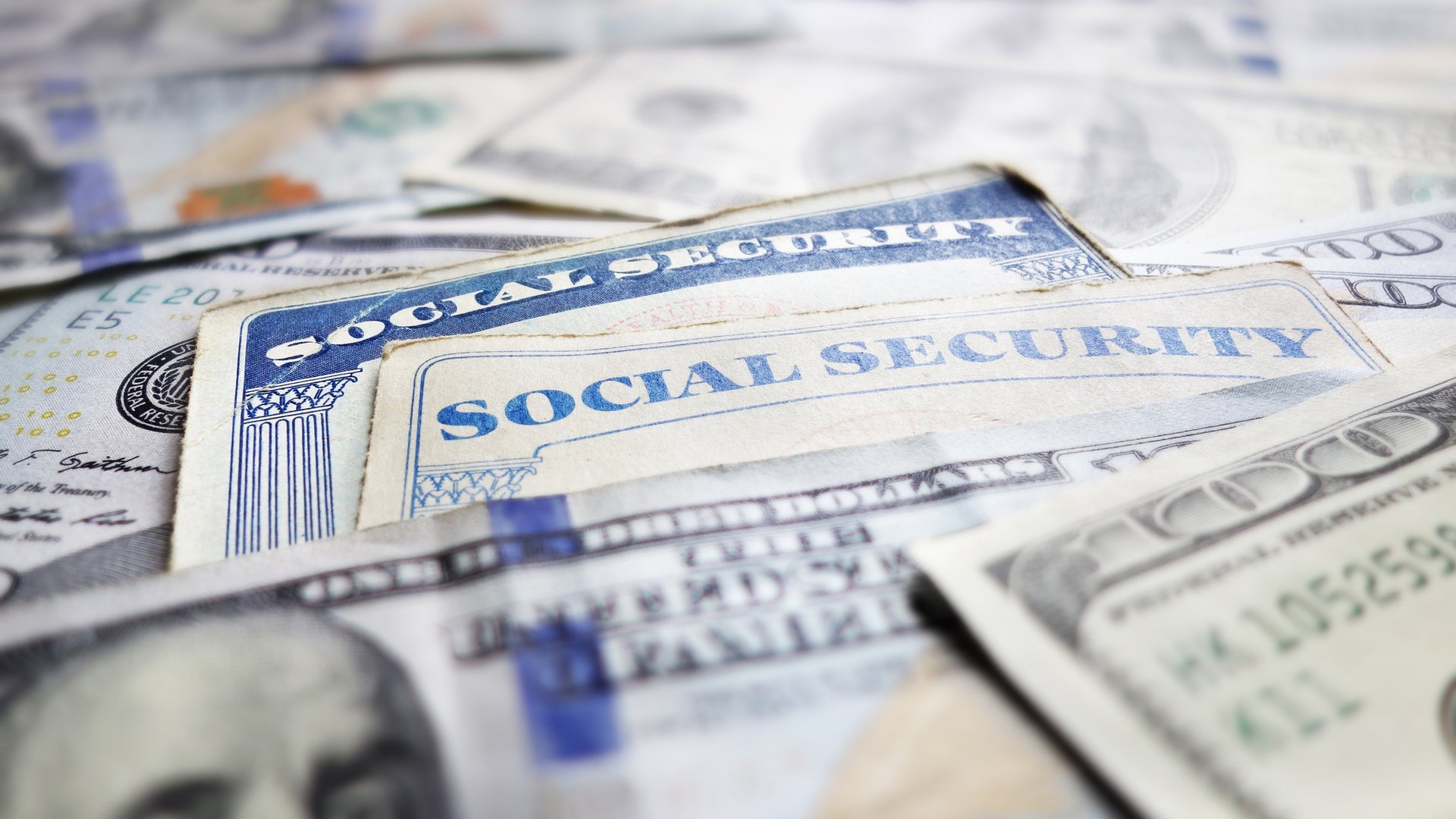 Discover two Social Security cards partially buried under a pile of hundred-dollar U.S. bills to showcase benefits.