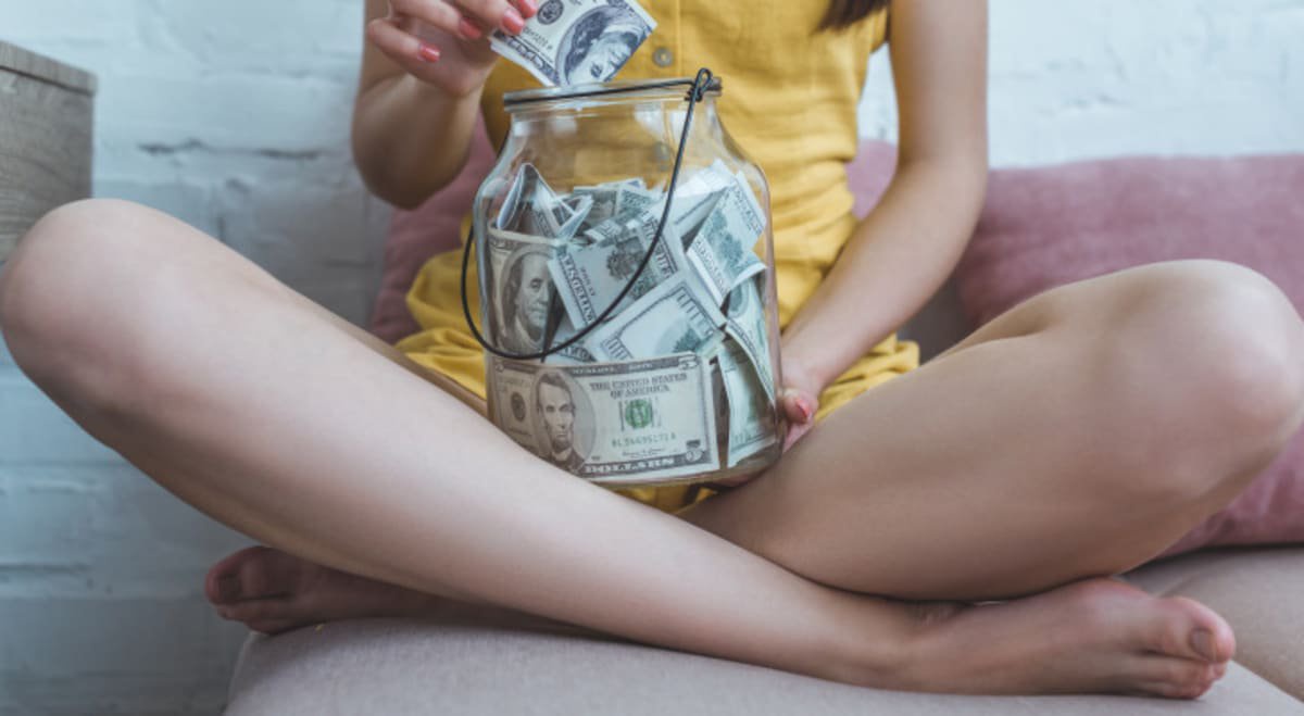 A young woman with braided brown hair is visible, sitting cross-legged with a glass jar full of American dollar bills in her lap.