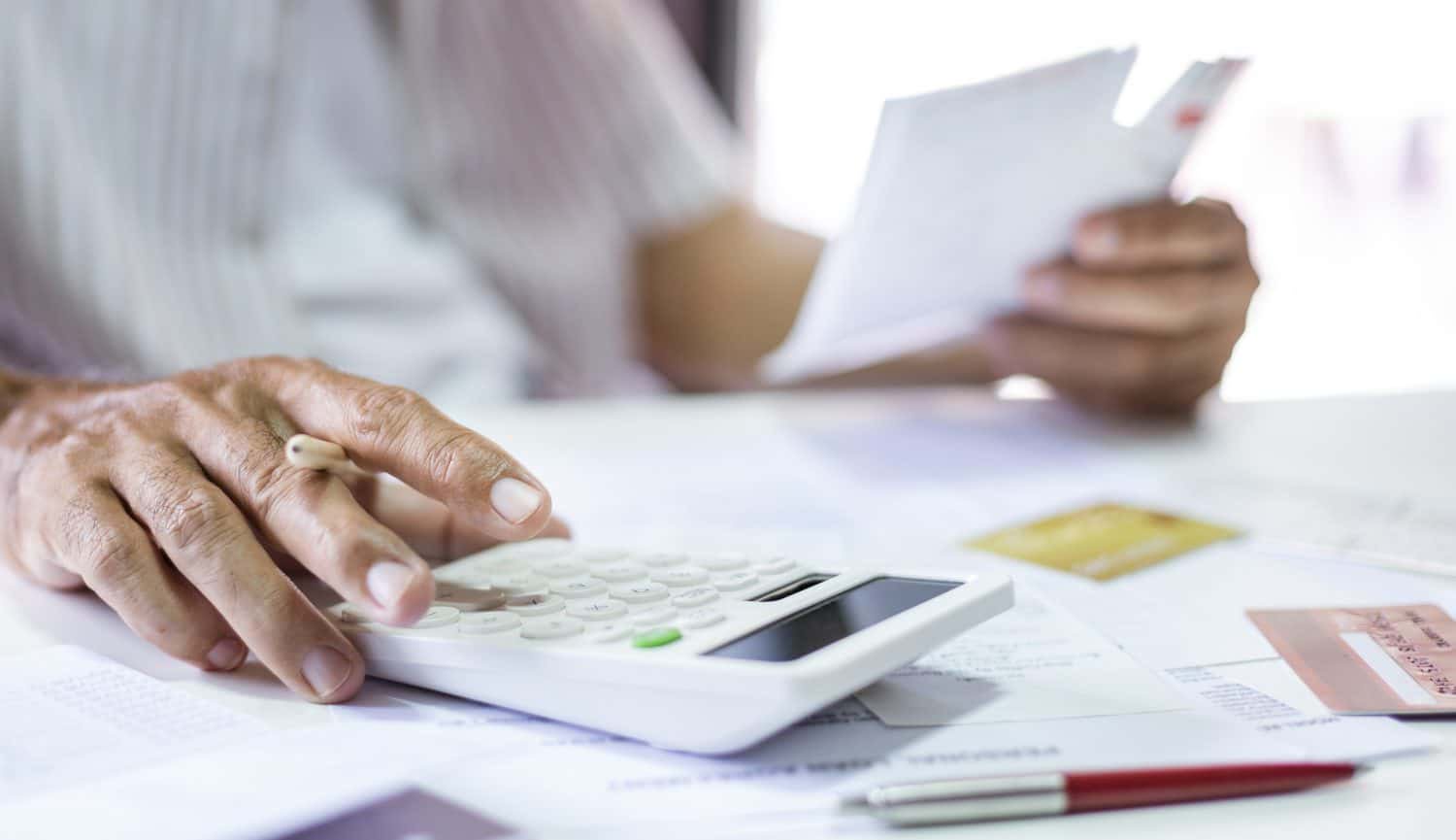 A close-up view shows a person's hands on a white desk. One hand, holding a pencil, presses buttons on a white calculator, while the other hand holds several white papers. Various financial documents, a red and silver pen, and several credit cards are spread across the light-colored surface.