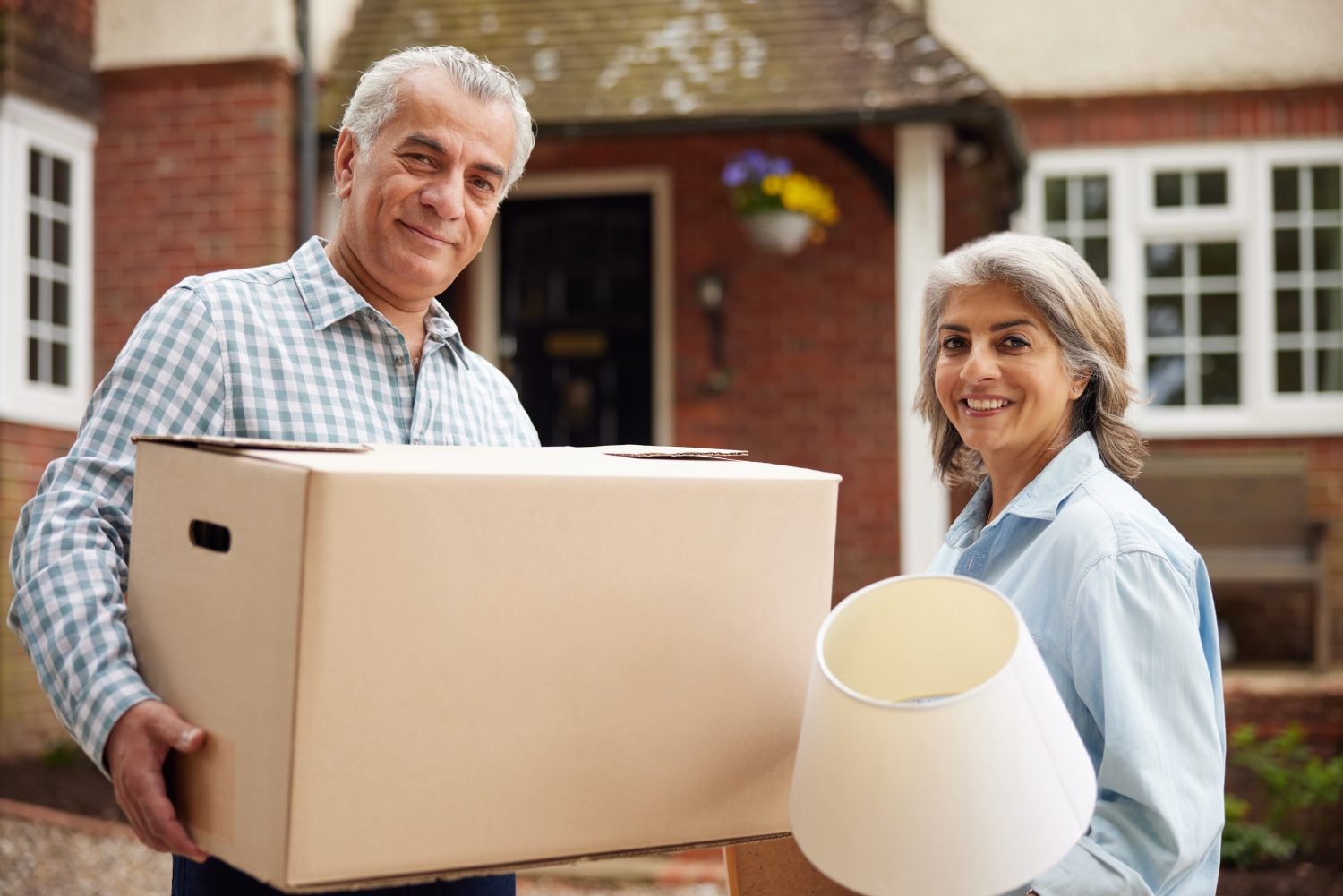 A smiling older man with gray hair in a plaid shirt holds a large cardboard moving box. Next to him, a smiling older woman with gray hair in a light blue shirt holds a white lampshade. They stand in front of a brick house with white windows, appearing to be in the process of moving.