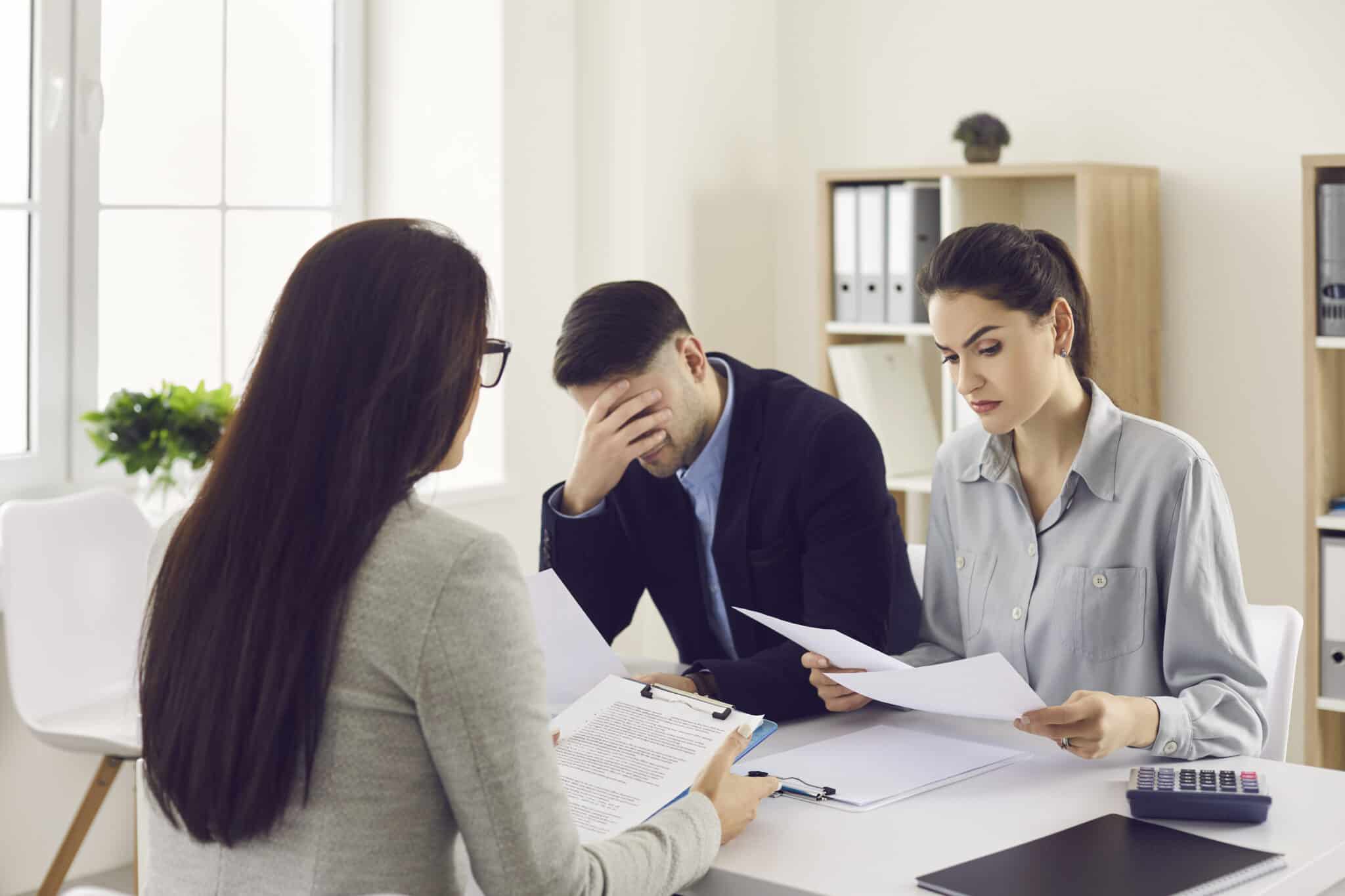 Three people sit around a white table. A woman with long dark hair, seen from behind, wears glasses and holds a clipboard. Across from her, a man in a dark suit covers his face with his hand, appearing distressed. Next to him, a woman with dark hair pulled back in a ponytail wears a light blue shirt and looks down at papers she holds. Several documents are spread on the table in a brightly lit office setting.