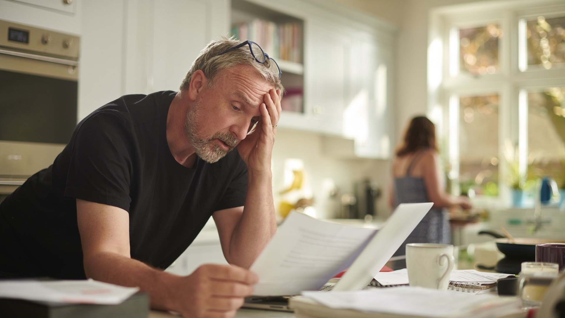 Discover a mature man looking stressed reviews financial paperwork with his spouse in the background