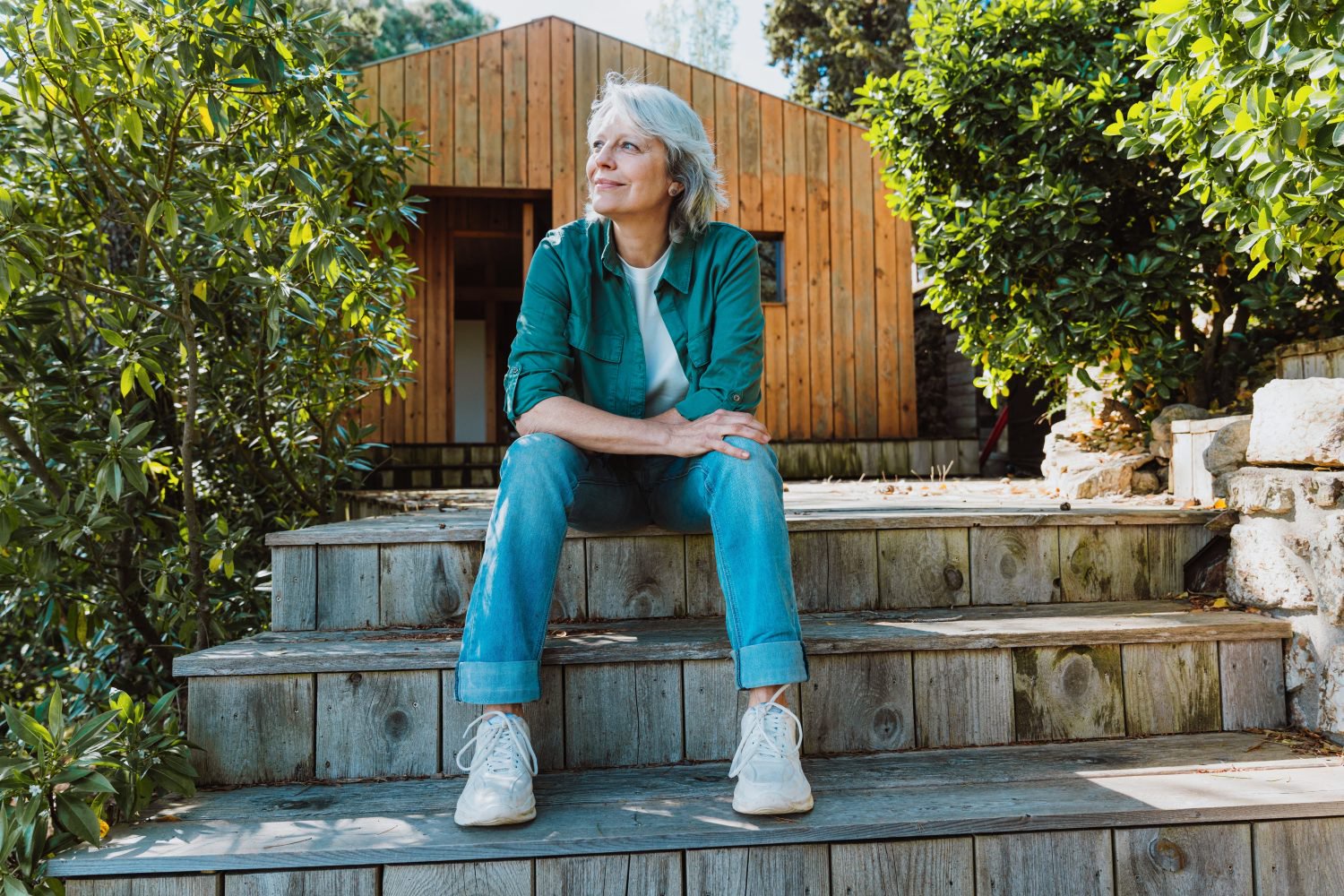 A smiling older woman with short gray hair, wearing a teal button-up shirt over a white tee, blue jeans, and white sneakers, sits relaxed on rustic wooden steps. Behind her is a light brown wooden cottage and lush green trees, bathed in bright sunlight.