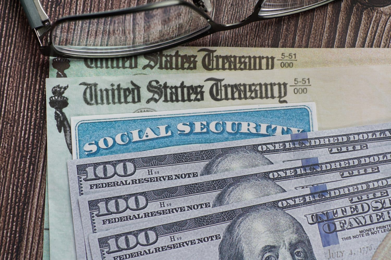 A pair of black-framed reading glasses rests atop several financial documents. Visible are two light green United States Treasury documents, a blue Social Security card, and multiple fanned-out one-hundred dollar bills featuring Benjamin Franklin's portrait. All items are arranged on a textured wooden surface.