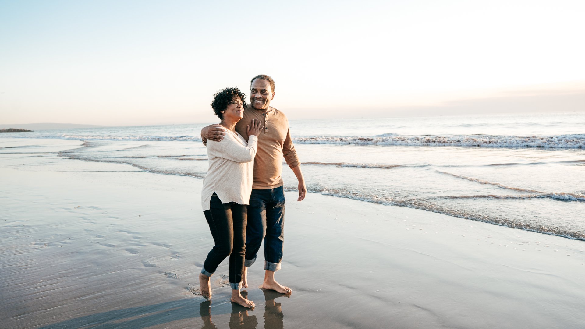 Discover a mature, retirement-aged couple smile with their arms around each other as they happy walk along the beach