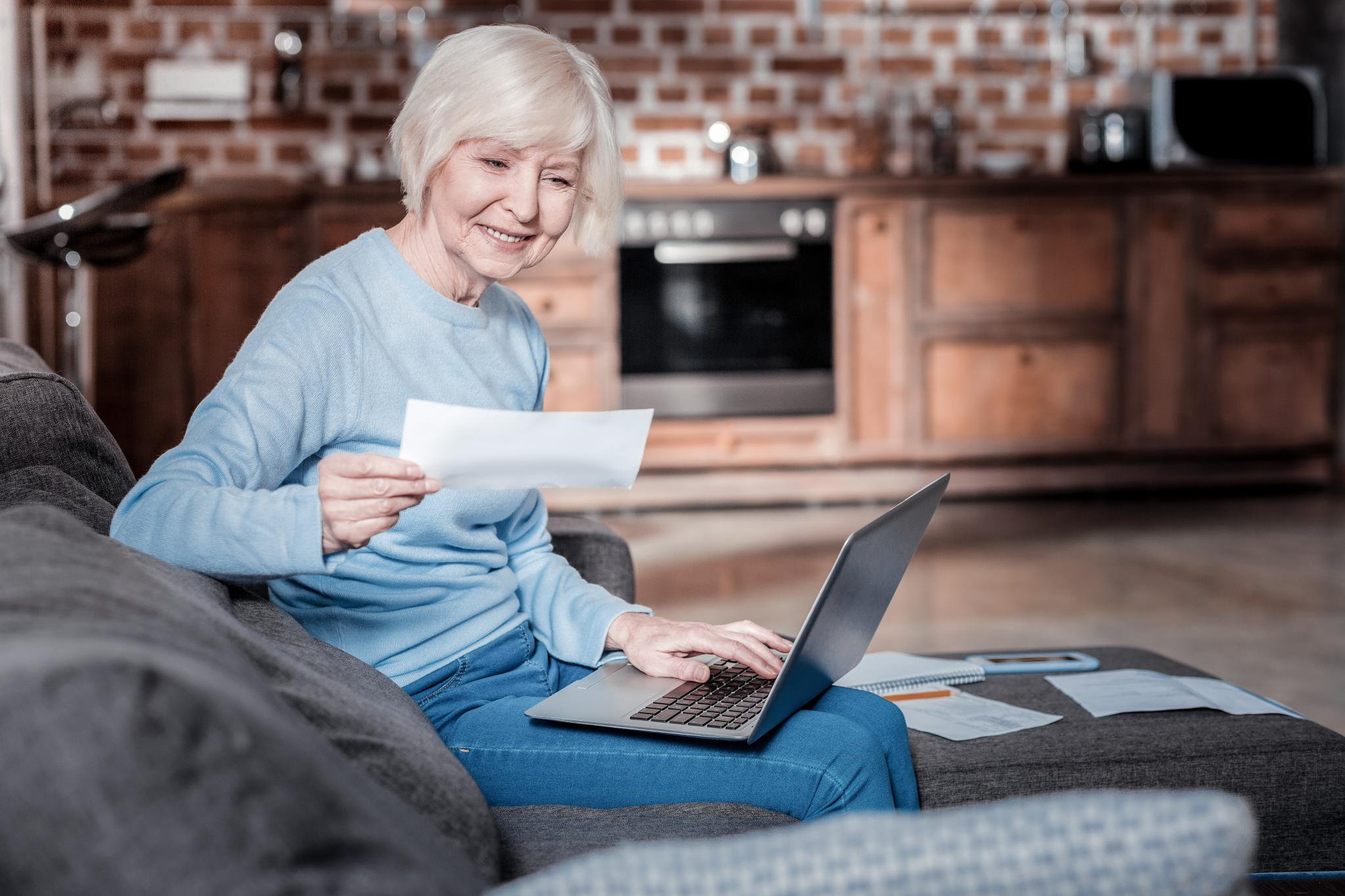 A smiling mature woman sits on a sofa with a laptop, reviewing documents and managing her finances at home.