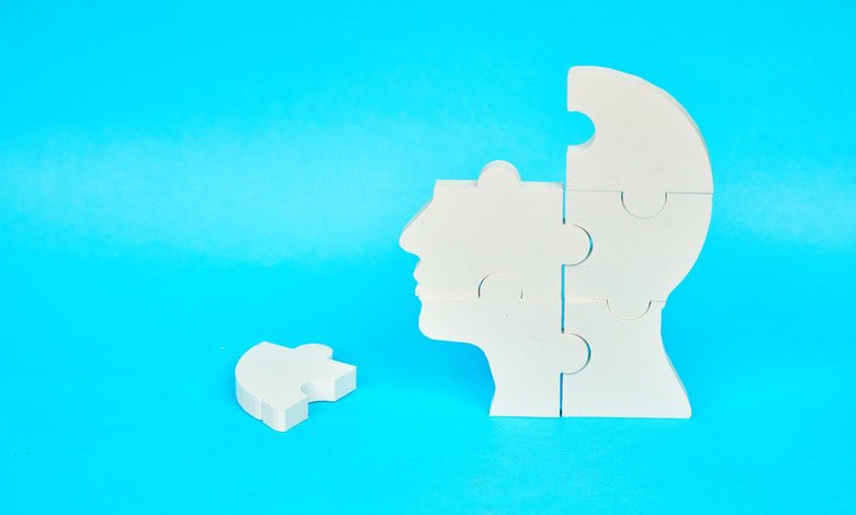 A white puzzle shaped like a human head with a missing piece resting beside it on a bright blue surface.