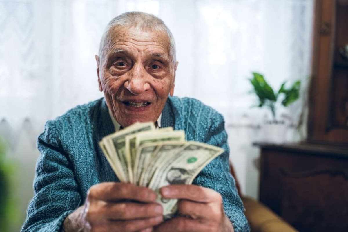 Smiling elderly man in a blue sweater holding a fan of US dollar bills in a cozy living room
