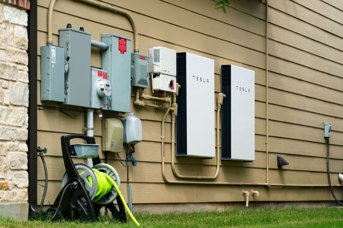 Two Tesla home battery units mounted on the side of a house near electrical equipment and a garden hose reel.