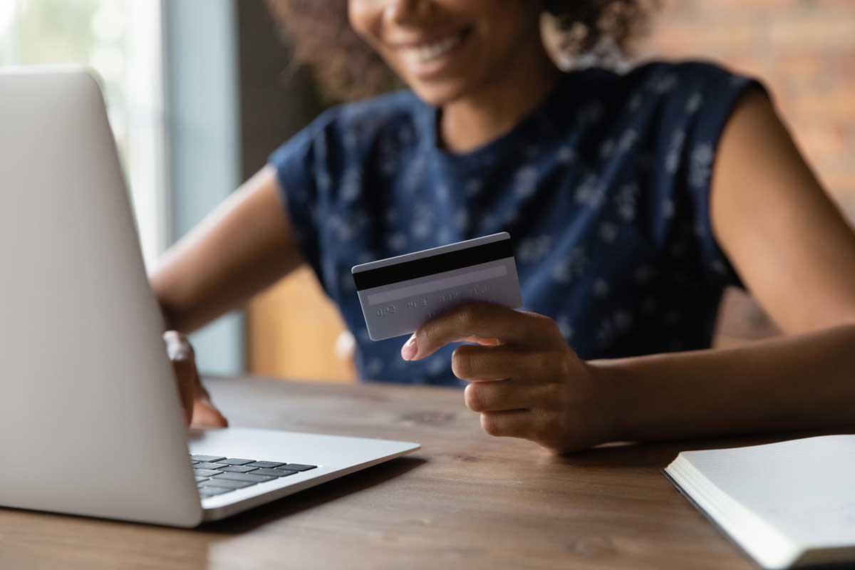 A person smiles while holding a credit card and using a laptop at a wooden desk, suggesting online shopping or banking. A notebook is partially visible next to the laptop.