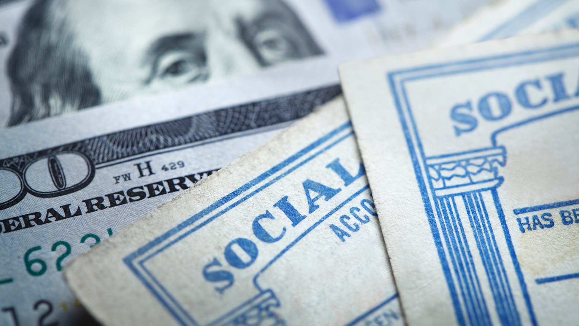 Discover two Social Security cards rest on top of several $100 bills with Ben Franklin peeking over them